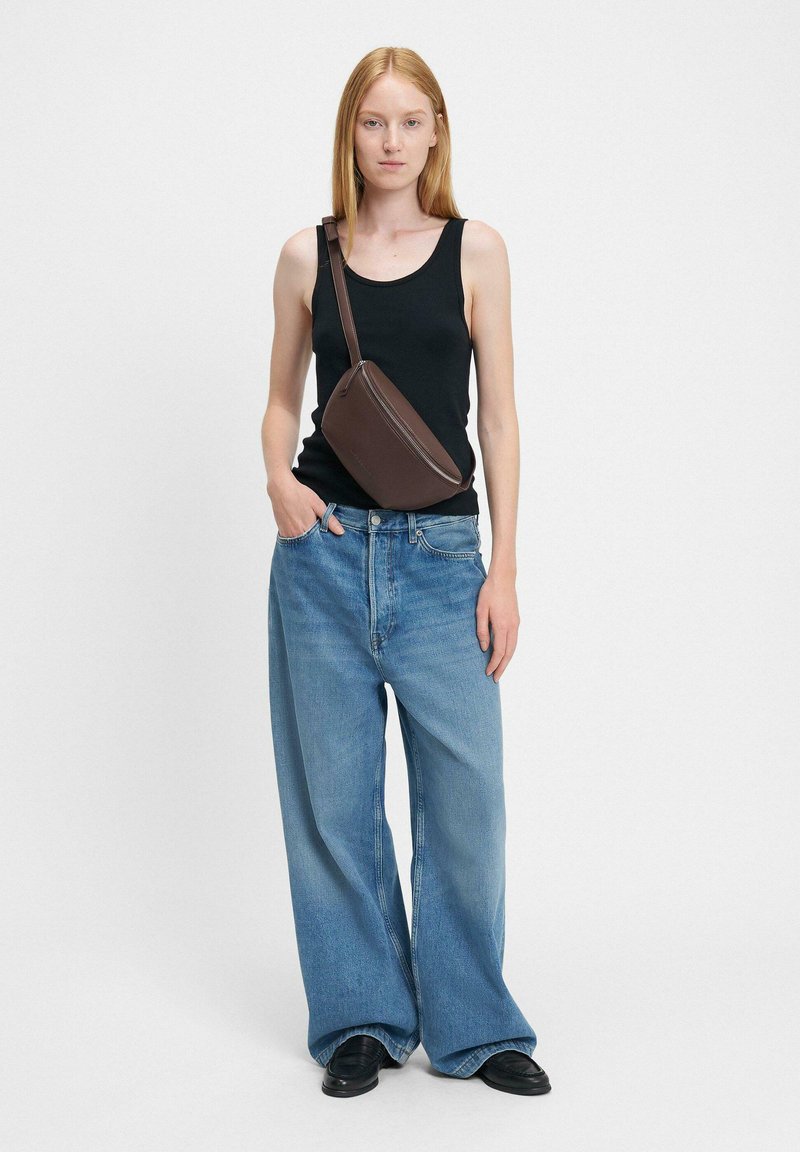 Borsa a spalla marrone con cerniera, indossata con canotta nera e jeans blu chiari larghi, abbinata a scarpe nere. Sfondo neutro.