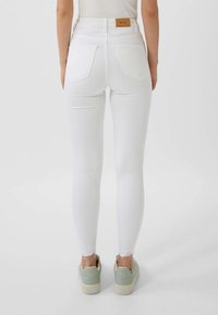 Los jeans blancos ajustados cuentan con una cintura alta, un sutil desgaste en el dobladillo y dos bolsillos traseros. Combinados con zapatillas verde claro.