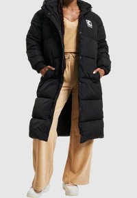 Černý péřový kabát s kapucí, zdobený prošívaným vzorem, logem na hrudi a postranními kapsami; nošený přes béžov�é kordové kalhoty.