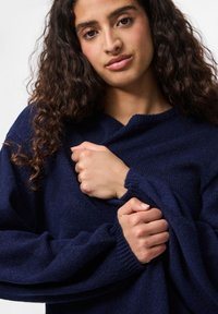 Sweater azul-marinho feito de tecido texturizado com um corte descontraído, apresentando mangas largas e um design de decote em V subtil.