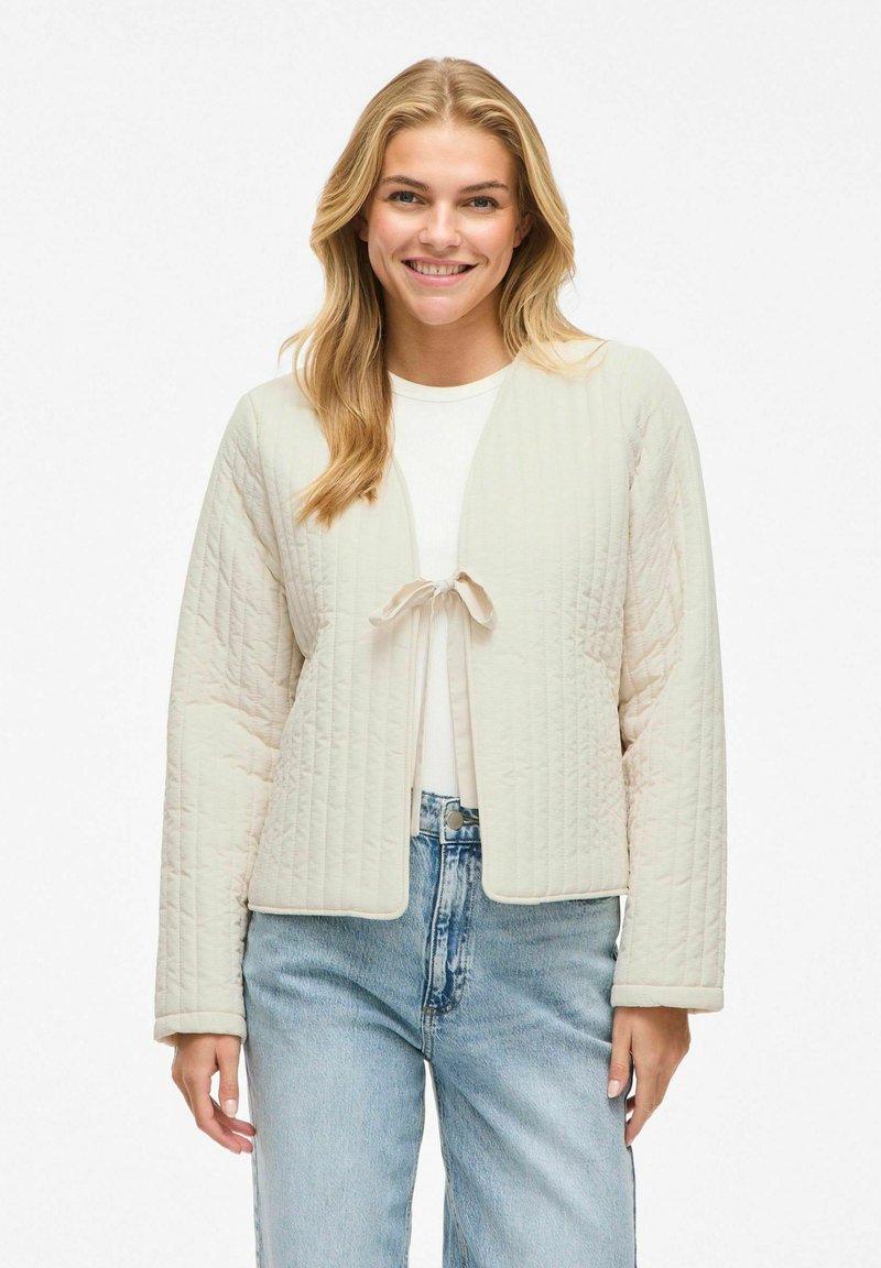 Giacca trapuntata in beige chiaro con design cropped, chiusura con lacci e maniche lunghe. Presenta un tessuto testurizzato e una vestibilità rilassata.
