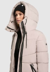 Svetloružová puffová bunda s veľkou kapucňou, čiernymi zipsami a sťahovacími šnúrkami. Má prešívaný dizajn a logo nášivku na rukáve.