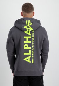 Sudadera gris con un gran gráfico vertical amarillo "ALPHA INDUSTRIES" en la parte posterior. Diseño con capucha, puños y dobladillo de canalé, con un bolsillo en la manga.