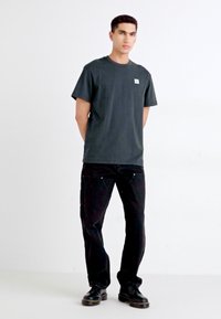 Maglietta in cotone grigio scuro con colletto a girocollo e piccolo logo sul petto, abbinata a pantaloni neri e scarpe nere, in piedi su uno sfondo bianco.