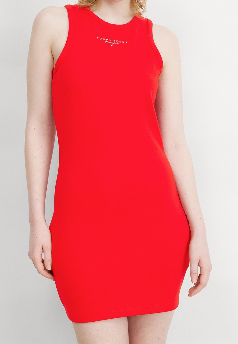 Femme portant une robe rouge ajustée sans manches avec le logo « Tommy Jeans New York » sur la poitrine, debout devant un fond uni.
