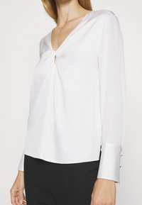 Bílá blůza s hlubokým výstřihem do V, uzlovým detailem vpředu, dlouhými průhlednými rukávy a knoflíkovými akcenty na manžetách. Hodvábná textura, volný střih.