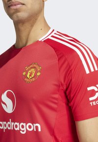 Maillot rouge de Manchester United en tissu texturé, avec des rayures blanches sur les épaules et le logo arboré sur la poitrine. Présente des logos de sponsors.