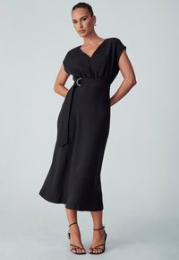 Mustat midi-mekko, jossa on lyhyet hihat, v-aukko, kapeat vyötärö ja vyö, sileä tekstuuri ja liehukehame. Tyylitelty mustilla remmi sandaaleilla.