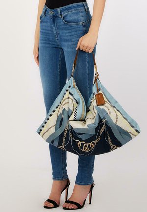 Donna che tiene una grande borsa di stoffa con motivo a catena blu, bianco e oro, indossa jeans skinny blu e sandali neri con tacco alto.