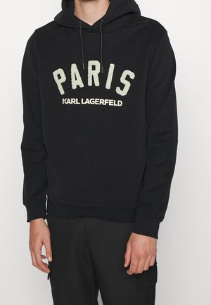 Czarna bluza z kapturem z "PARIS" dużymi białymi literami oraz "KARL LAGERFELD" mniejszym białym tekstem poniżej, noszona przez osobę.