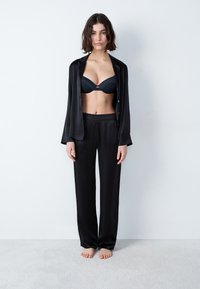 Zwarte satijnen langemouwshirt over een zwarte bh en bijpassende broek, met een slank ontwerp en een soepele textuur. Model staat blootsvoets.