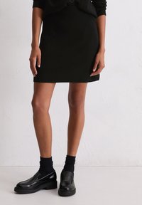 Jupe en tricot noire au design ajusté, longueur mi-cuisse, avec des coutures latérales discrètes. Associée à des chaussures noires à enfiler et des chaussettes jusqu'à la cheville.