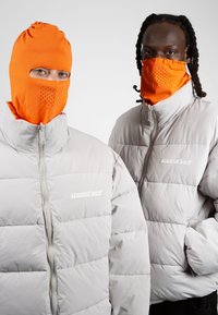 Chaquetas acolchadas de color gris claro con un cuello alto, con un logo. Mascarillas naranjas con detalles perforados, usadas por dos personas.