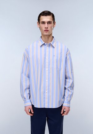 Mladý muž stojící, oblečený ve světle modré a béžové košili s vertikálními pruhy a tmavě modrých kalhotách, čelící dopředu na jednobarevném pozadí.