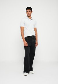 Fehér galléros póló, fekete bő szárú farmernadrág és fehér slip-on cipő. A modell karjai lazán pihennek, egyszínű világos háttér előtt áll.