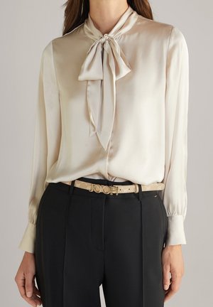 Femme portant un chemisier en satin crème avec un col noué en nœud papillon, un pantalon noir taille haute et une ceinture beige avec une boucle dorée de marque.