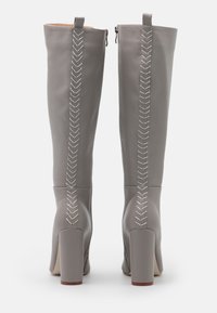 Un par de altas botas de cuero gris con tacón vistas desde atrás, con costuras decorativas blancas a lo largo del centro.