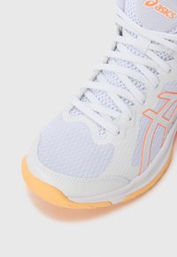 Hvit idrettssko med et teksturert mesh-overdel, oransje detaljer og en lys oransje gummisåle. Har et klassisk snøringsdesign.