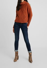 Tjock orange stickad tröja med munkframskärning, med diamantmönster; matchad med mörka smala jeans och ankelboots med ormtryck.