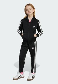 Ung tjej som står i en svart Adidas-träningsdräkt med vita ränder, händerna i fickorna, iförd vita Adidas-sneakers och rosa strumpor.