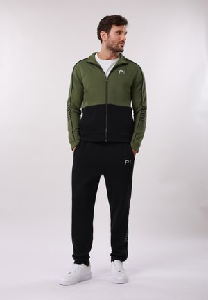 Uomo che indossa una giacca con cerniera verde e nera e pantaloni da jogging neri con scarpe da ginnastica bianche, in piedi con le mani nelle tasche, guarda verso destra.