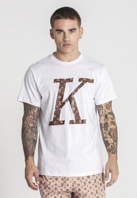 Bílý bavlněný tričko se velkým hnědým grafickým písmene "K" se vzorem log. Krátké rukávy a klasický kulatý výstřih.