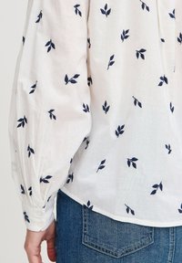 Weiße Bluse mit marineblauem Blattstickerei, Puffärmel und Knopfmanschetten. Weicher, leichter Stoff und lockere Passform.