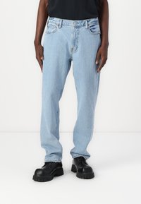Jeans de mezclilla azul claro con corte recto, que cuentan con bolsillos frontales, passacintos y una textura desgastada, combinados con zapatos negros de suela gruesa.