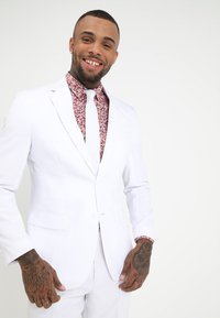 Traje blanco con solapas afiladas, usado sobre una camisa de patrón floral rosa y corbata blanca. Las características clave incluyen intrincados patrones de flores y manos tatuadas.