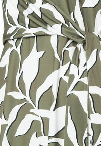 Grüner Stoff mit einem weißen Blumenmuster, das von Schwarz umrandet ist; zeigt ein geknotetes Detail, wo der Stoff sich überlappt und dadurch Textur und Tiefe erzeugt.