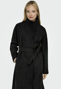 Manteau noir long avec un col haut et une taille ceinturée. Présente une texture douce et des poches latérales ; les manches sont amples et légèrement évasées.