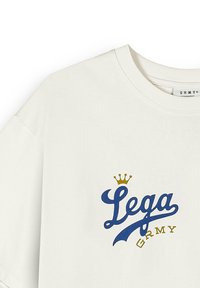 Kremowa bawełniana koszulka z krótkim rękawem, z niebieskim i złotym wzorem graficznym, na którym znajduje się napis "Lega GRMY" oraz korona.