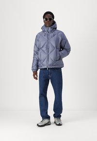 Chaqueta gris acolchada con capucha y patrón de diamantes, combinada con jeans azules y zapatos grises y negros. El modelo lleva gafas de sol oscuras.