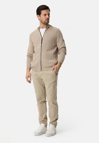 Beige zip-up cardigan with a shawl collar and ribbed cuffs, paired with light tan pants and white sneakers, set against a plain background.