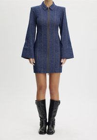 Denimminiklänning med långa klockärmar, dragkedja framtill och kontrasterande sömmar. Bärs med knähöga svarta läderstövlar.