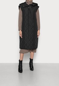 Chaleco negro acolchado sin mangas con capucha, con botones a presión y un bolsillo, usado sobre un suéter de cuello alto marrón y una falda beige.