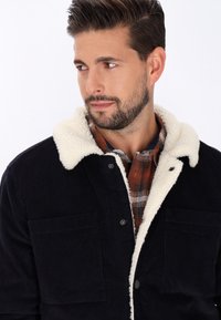 Marineblaue Cordjacke mit einem nachtblauen Fleece-gefütterten Kragen, ausgestattet mit zwei Brusttaschen und einer Knopfverschluss, getragen über einem karierten Hemd.
