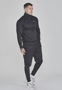 Chándal deportivo negro con una chaqueta con cremallera y pantalones jogger, que cuenta con puños acanalados, bolsillos laterales y acentos de logo en blanco contrastante.
