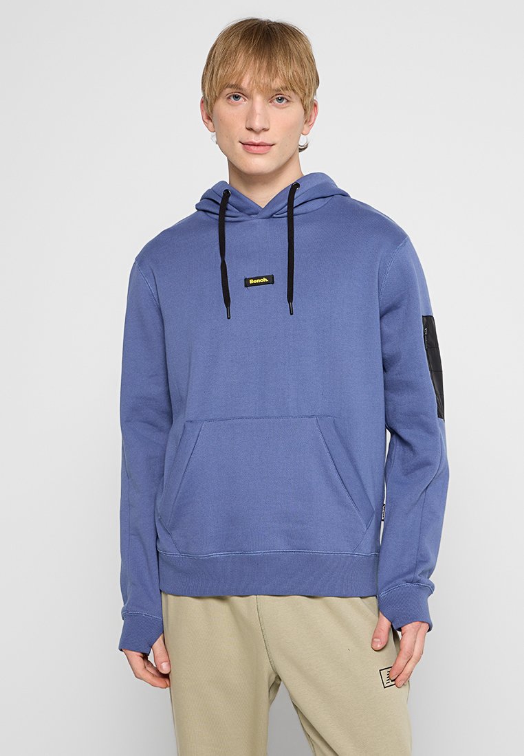 Joven con cabello corto rubio que lleva una sudadera azul con capucha y cordones negros, y pantalones beige, de pie frente a un fondo simple.