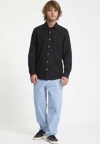 Chemise noire à manches longues boutonnée avec une poche poitrine, portée avec un jean bleu clair ample et des baskets noires.