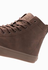 Zapato alto sintético marrón con punta redonda, superficie texturizada, seis ojetes y suela gruesa. Presenta un logo sutil en el costado.