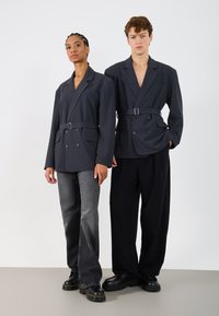 Două modele poartă sacouri supradimensionate, de culoare gri închis, cu dublu sucol, prevăzute cu curele, asortate cu pantaloni negri și gri și cizme voluminoase negre.