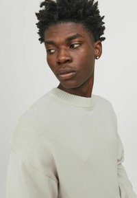 Light gray, knitted sweater with a ribbed crew neckline and dropped shoulder seams, worn by a person with short, textured hair.