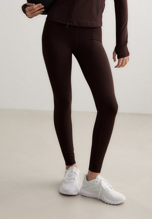 Person iført mørkebrune leggings, matchende jakke og hvide atletiske sneakers, der står på en lys gulv mod en ensfarvet væg.