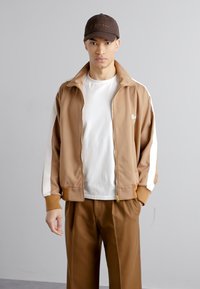 Giacca a track marrone con strisce bianche laterali, zip frontale, colletto a coste, indossata sopra una maglietta bianca e pantaloni marroni. Cappellino marrone con logo.