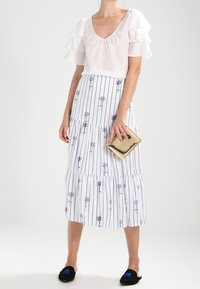 Žena v bílé blůze s volány na rukávech, bílé midi sukni s modrými pruhy a květinovým potiskem, držící zlatou kabelku a oblečená v černých slip-on botách.