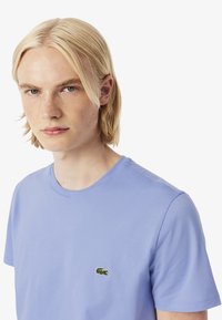 Een jonge persoon met blond haar draagt een lichtblauw T-shirt met een klein groen Lacoste-logo op de borst. De achtergrond is eenvoudig wit.