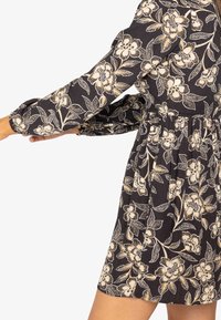 Vestido negro con flores beige, mangas largas abullonadas y cintura ajustada. Presenta una tela texturizada y una falda acampanada.