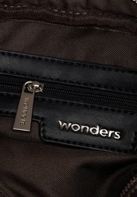 Primer plano de un tirador de cremallera de cuero negro y una etiqueta con la marca plateada "wonders" sobre un fondo de tela marrón oscuro.