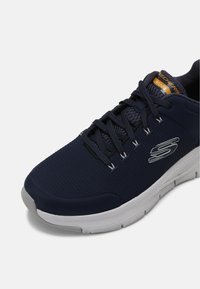 Zapatilla deportiva azul marino con parte superior de malla, suela de goma blanca y acentos grises. Presenta cordones y etiqueta 'Skechers Arch Fit'.